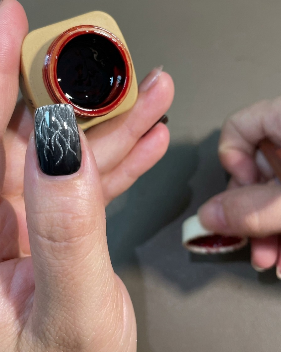 Ein Nail-Art-Pinsel trägt ein transparentes, dunkelrotes Gel über ein ausgehärtetes schwarz-weißes Cat-Eye-Flammendesign auf.