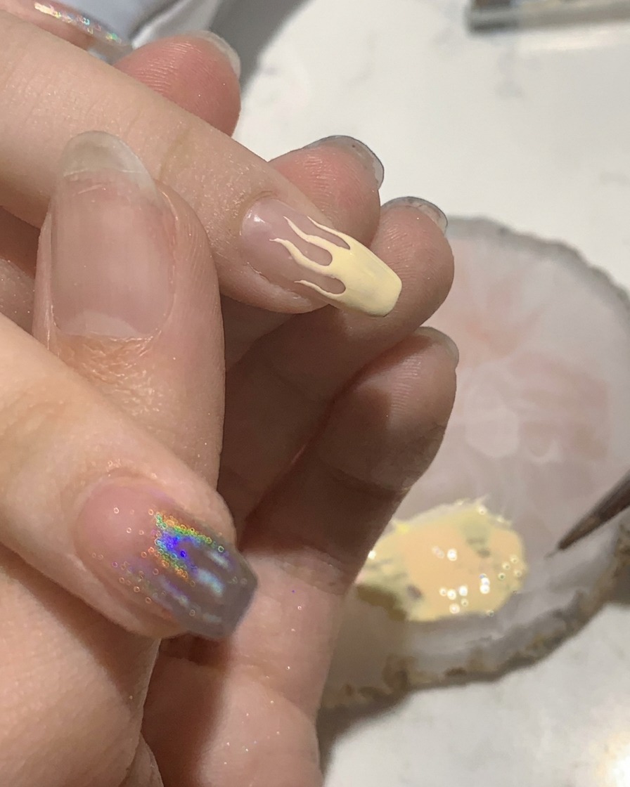 Ein kleiner flacher Pinsel füllt ein pfirsichfarbenes Flammen-Design auf einem Fingernagel aus.