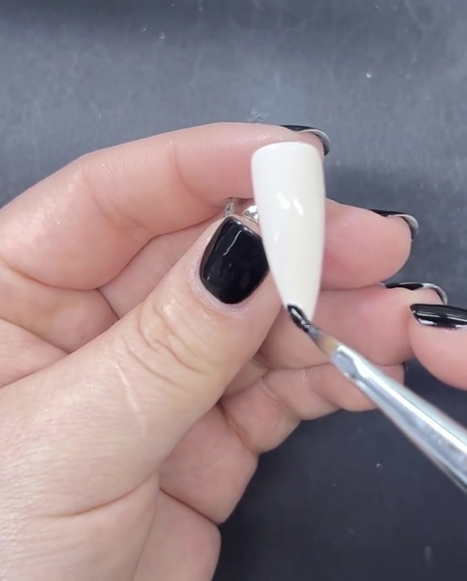 Ein feiner Liner-Pinsel trägt eine schmale, präzise Linie schwarzes Gel auf die Spitze eines milchig-weiß lackierten Nagels auf.