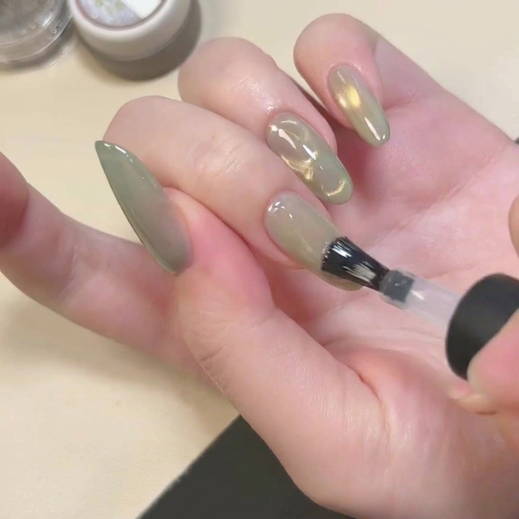 Ein Pinsel trägt einen klaren, matten Überlack auf den grünen Cat Eye Nagel auf.