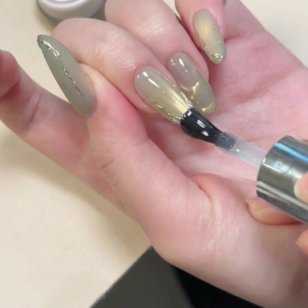 Ein Pinsel trägt glänzenden Überlack über den matten Nagel mit goldenen Akzenten auf.