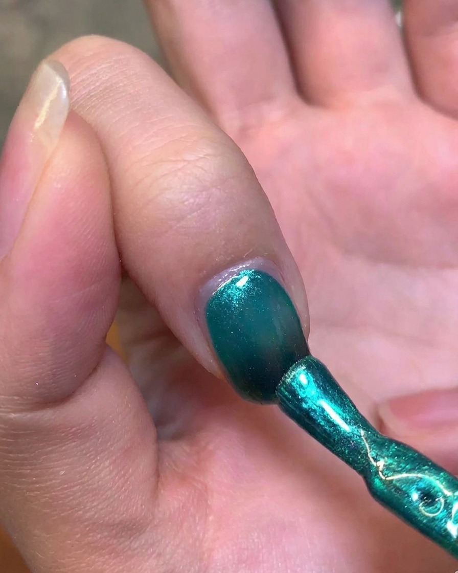 Ein Magnetstift aus Metall wird an die Seite eines Fingernagels gehalten, um das grüne Gel zu einem Cat-Eye-Highlight zu formen.