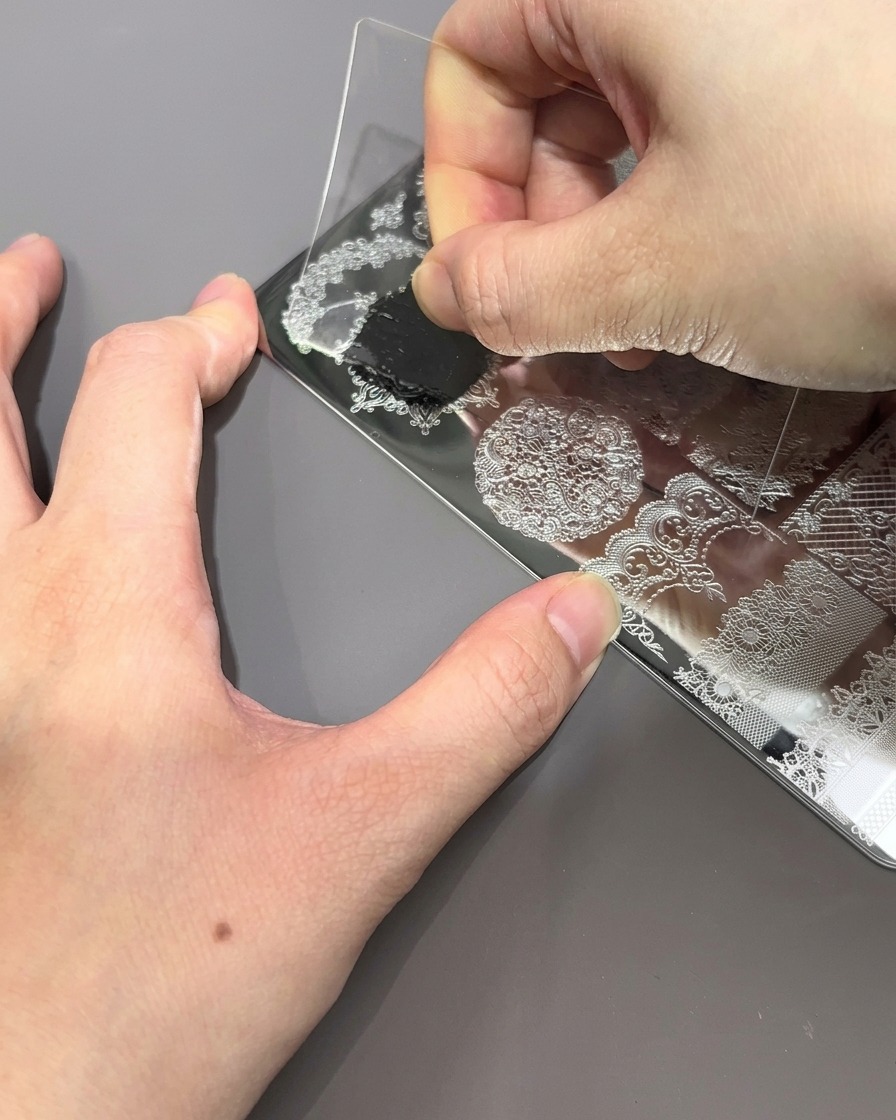Eine klare Kunststoffkarte wird über die Stamping-Platte gezogen und entfernt überschüssigen schwarzen Lack, wobei das gravierte Muster sichtbar wird.