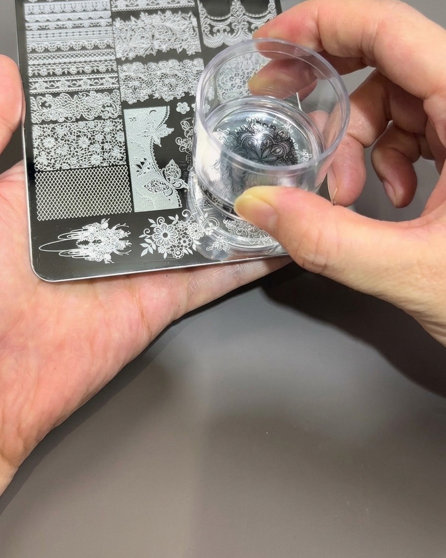 Eine Hand hält einen klaren Silikon-Nagelstempel und drückt ihn sanft auf das schwarze Motiv der Metallplatte.