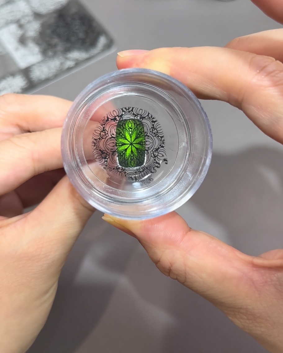 Blick durch einen klaren Silikonstempel, der zeigt, wie ein schwarzes Fenstergitter-Muster perfekt über einem grünen Cat-Eye-Nagel ausgerichtet ist.