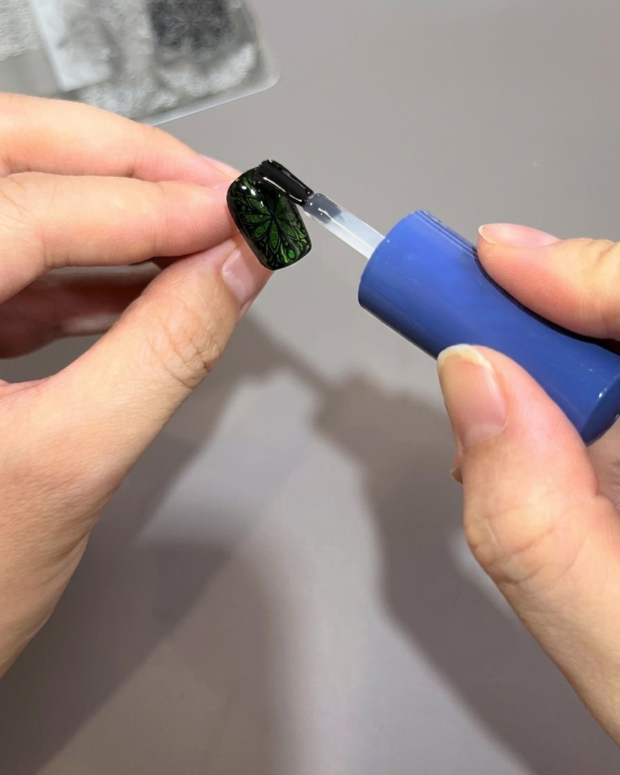 Ein Pinsel trägt eine dicke Schicht klaren, glänzenden Top Coat auf einen grünen Nagel mit schwarzem, gestempeltem Muster auf.