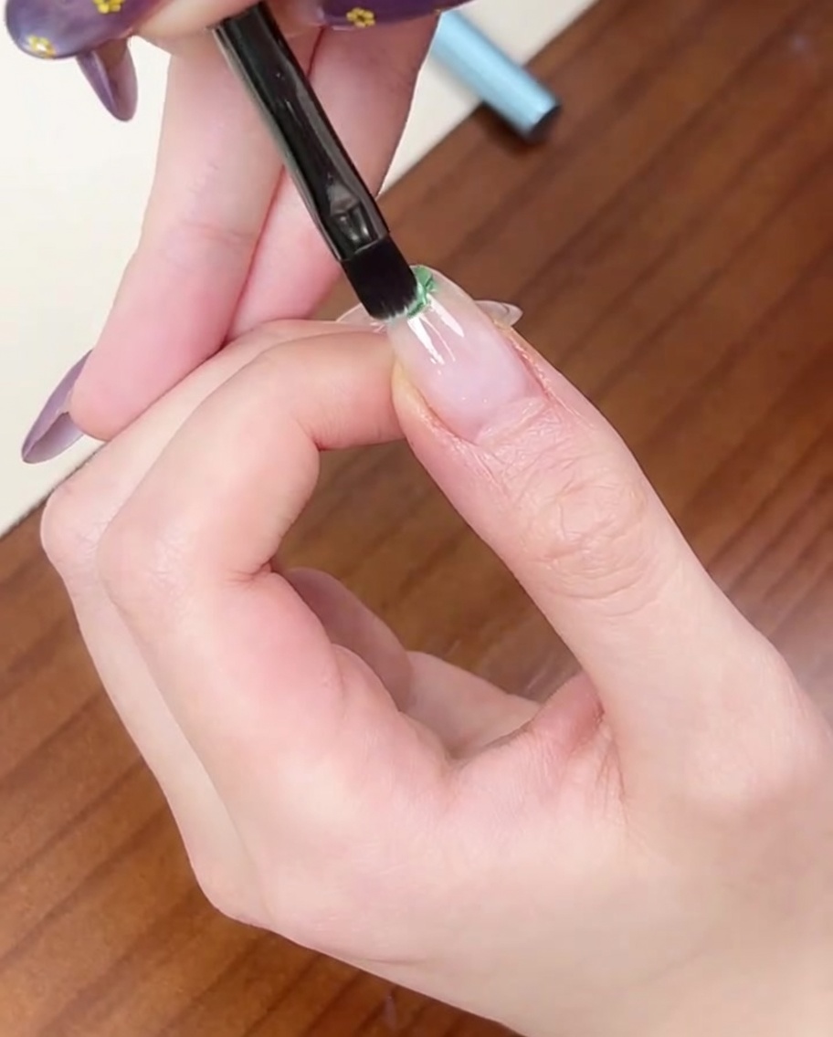 Ein flacher Pinsel trägt einen kleinen Tropfen hellgrünen Gellack auf die Spitze eines Naturnagels auf.