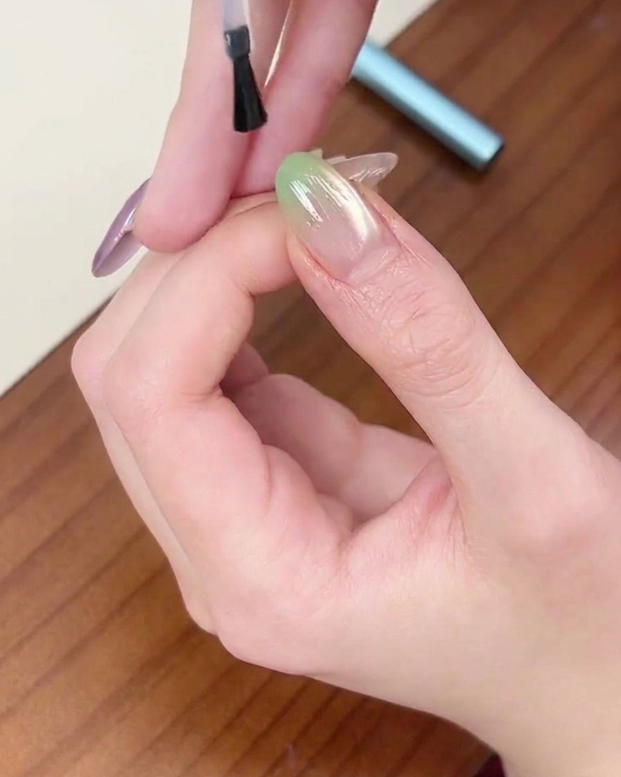 Ein Pinsel trägt eine abschließende Schicht klaren Top Coats über einen Nagel mit grünem Verlauf auf.