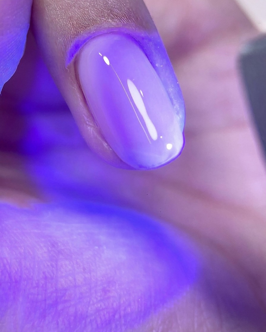Ein Fingernagel mit einer ungehärteten Schicht glänzendem rosa Gel unter dem leuchtend violetten Licht einer UV/LED-Nagellampe.