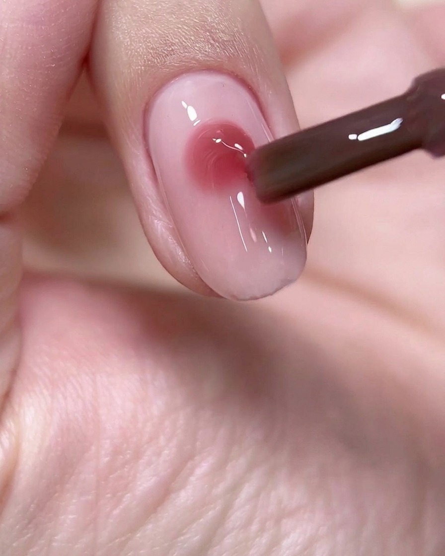 Ein Nagelpinsel, der einen präzisen, runden Tropfen tiefen weinrosa Gels in die Mitte einer hellrosa Nagelbasis setzt.