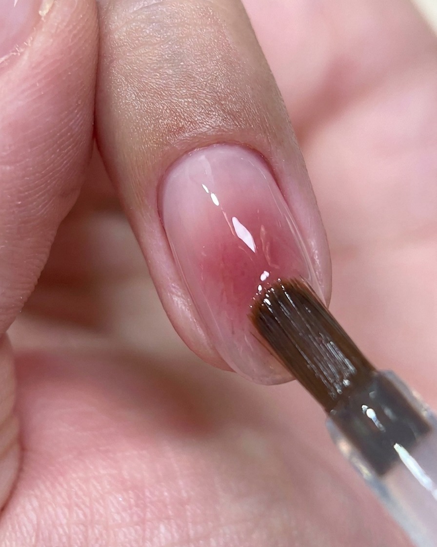 Ein Nagelpinsel setzt einen kleinen Tropfen klaren Top Coat Gels in die Mitte eines glänzenden rosa Nagels.