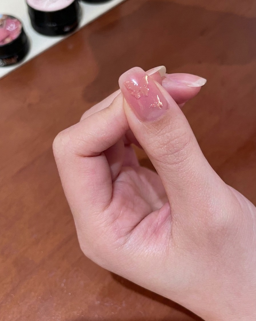 Eine Nahaufnahme einer Hand, die einen kleinen zylindrischen Magneten über einen mit feuchtem goldenen Magnetgel beschichteten Fingernagel hält und einen changierenden Schimmer enthüllt.