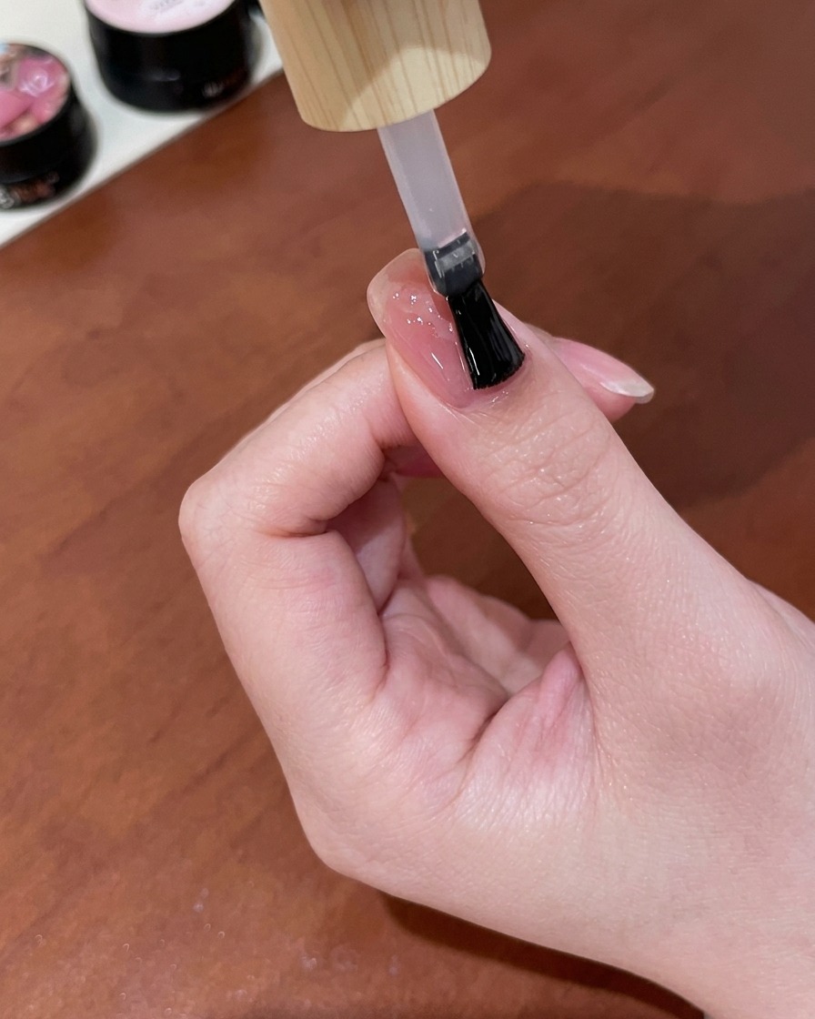 Ein Pinsel trägt eine Schicht klaren, glänzenden Gel-Top-Coat über einem fertigen nude-pinken Nagel mit einem goldenen Cat-Eye-Schmetterlingsdesign auf.