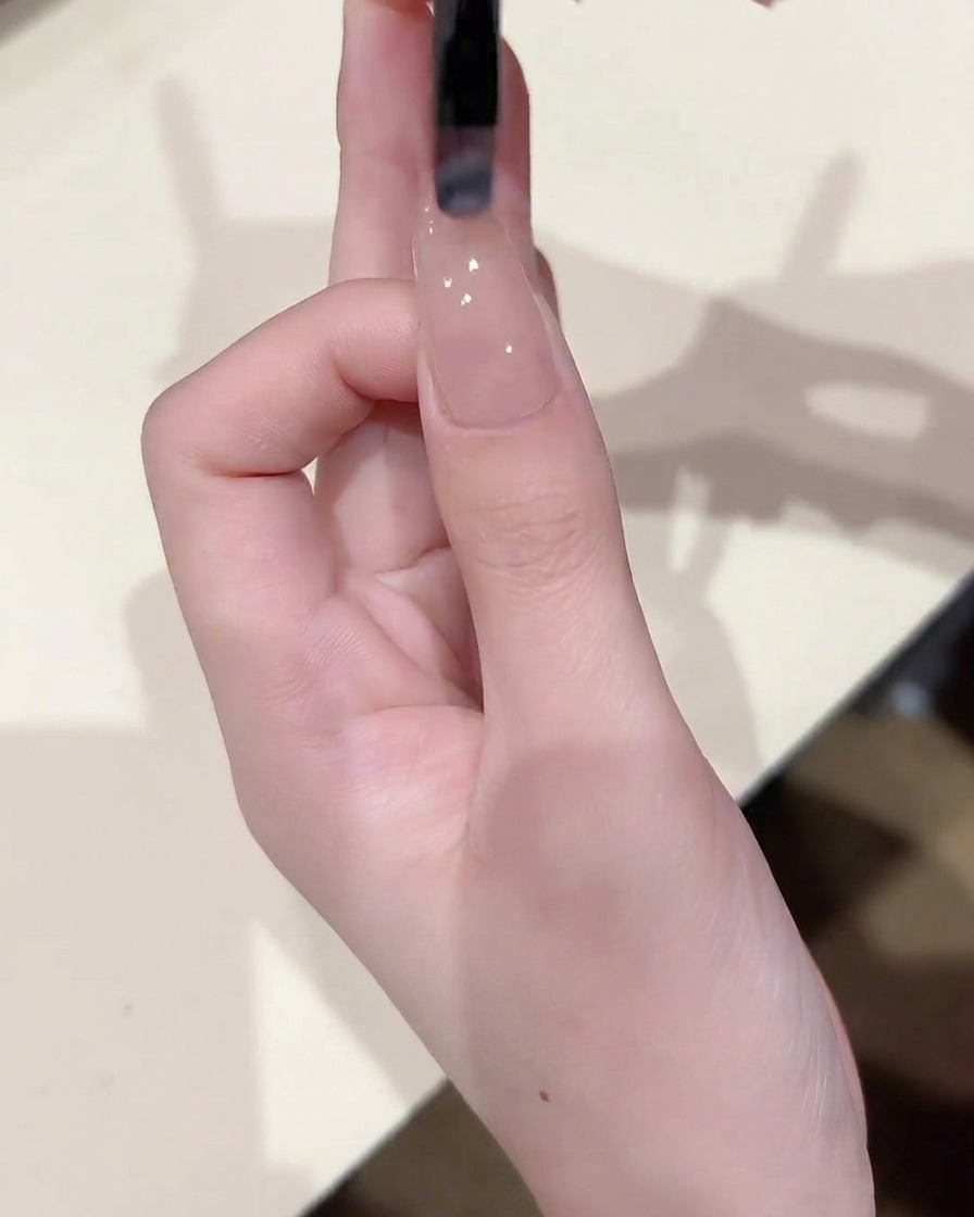 Nahaufnahme eines Pinsels, der eine dünne, gleichmäßige Schicht transparentes Nude-Gel auf den Nagel aufträgt.