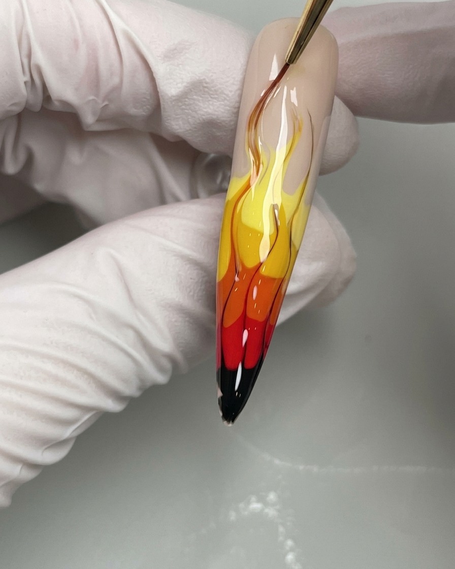 Nahaufnahme eines Nagelpinsels, der dünne schwarze Gellinien zieht, um die Ränder des gelben, orangen und roten Flammendesigns zu definieren.