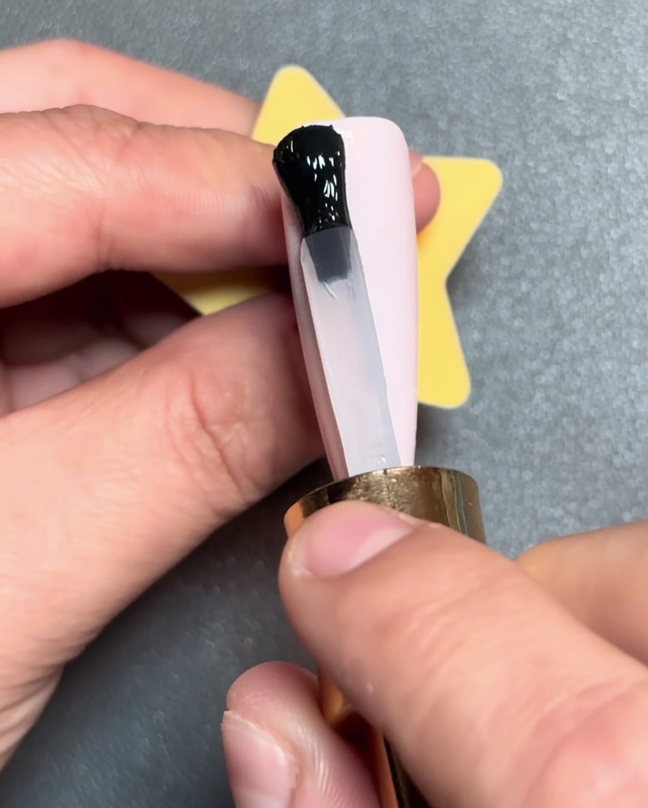 Ein Pinsel zieht eine horizontale schwarze Linie aus Gellack über die Spitze eines rosa Nagels.