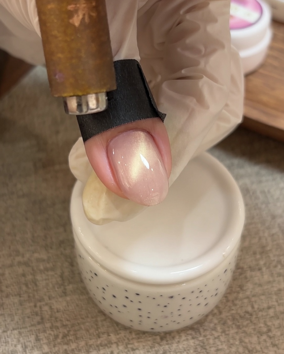 Eine Hand nutzt ein magnetisches Werkzeug, um die schimmernden Partikel zur Nagelspitze zu ziehen.