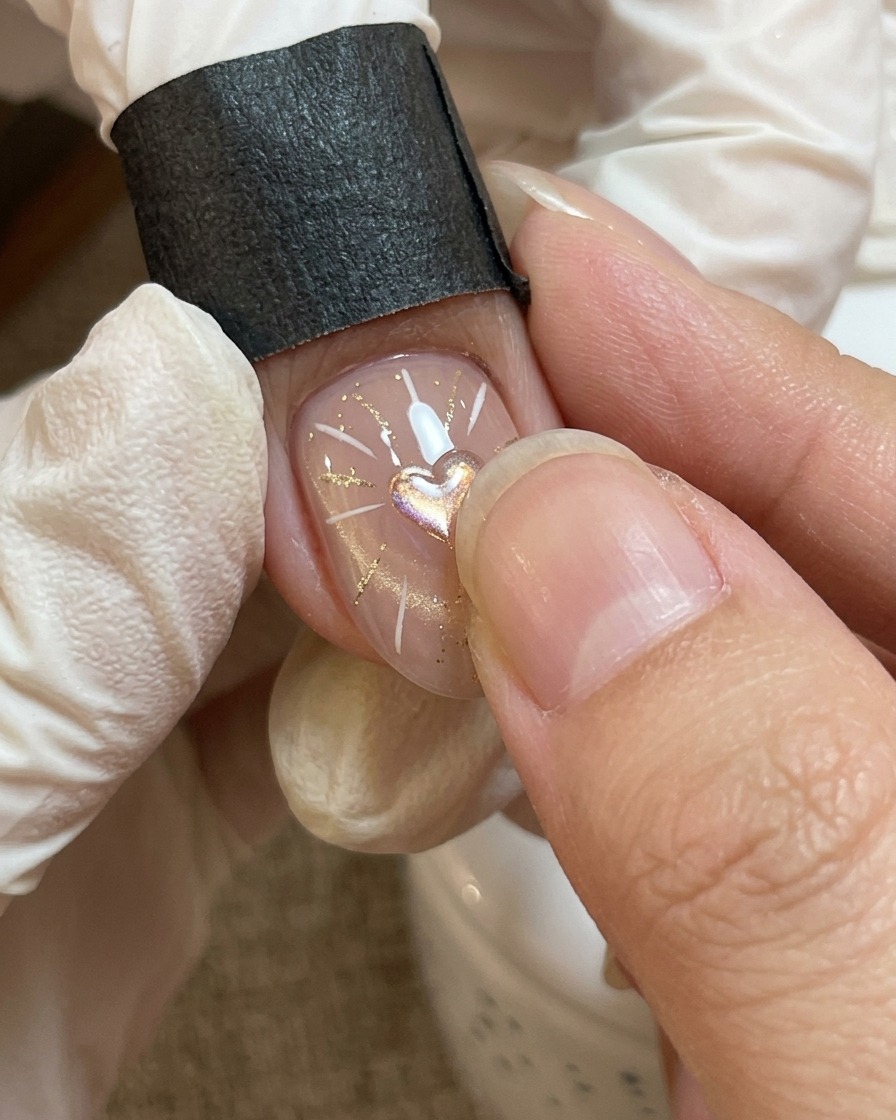 Nahaufnahme einer Nagelmodellage, bei der ein dickflüssiges, klares Gel vorsichtig über ein zentrales Herz-Motiv platziert wird.