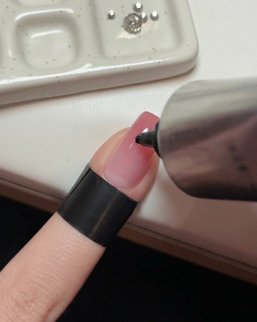 Eine Hand drückt einen Tropfen klares Klebegel auf die Mitte eines glänzenden rosa Nagels.