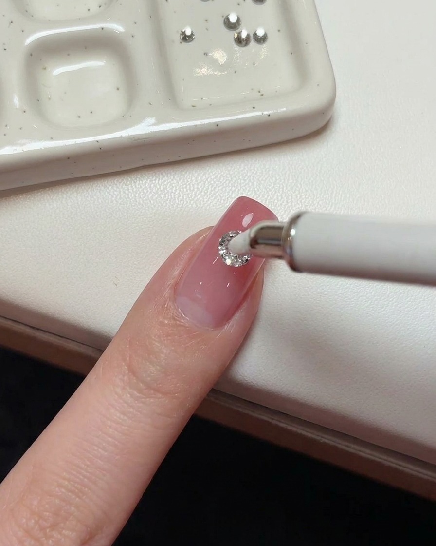 Ein Wachsstift platziert einen 5-mm-Kristall mittig auf dem rosa Nagel.