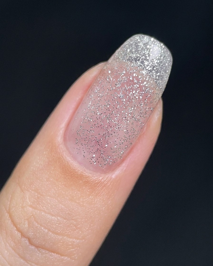 Ein Fingernagel, bedeckt mit einer dünnen, transparenten Schicht aus feinem Silber-Glitzergel.