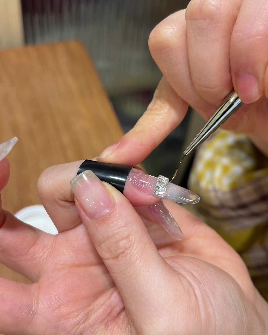 Nahaufnahme eines feinen Nail-Art-Pinsels, der hartes Top-Gel um die Basis eines silbernen Strass-Charms aufträgt.