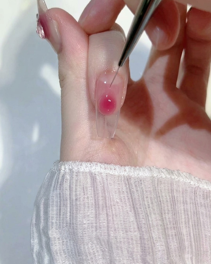Nahaufnahme eines feinen Liner-Pinsels, der einen Tropfen rosa Blush-Gel auf einer klaren Nagelverlängerung sanft verblendet, um einen weichen Farbverlauf zu kreieren.