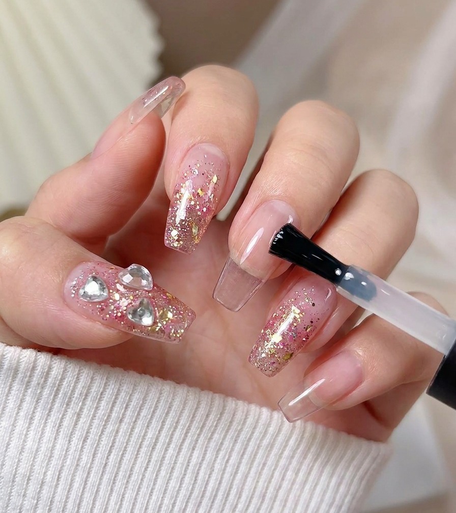 Ein Pinsel trägt einen abschließenden glänzenden Top Coat auf einen Nagel auf, der mit rosa Glitzer und Herzkristallen verziert ist.