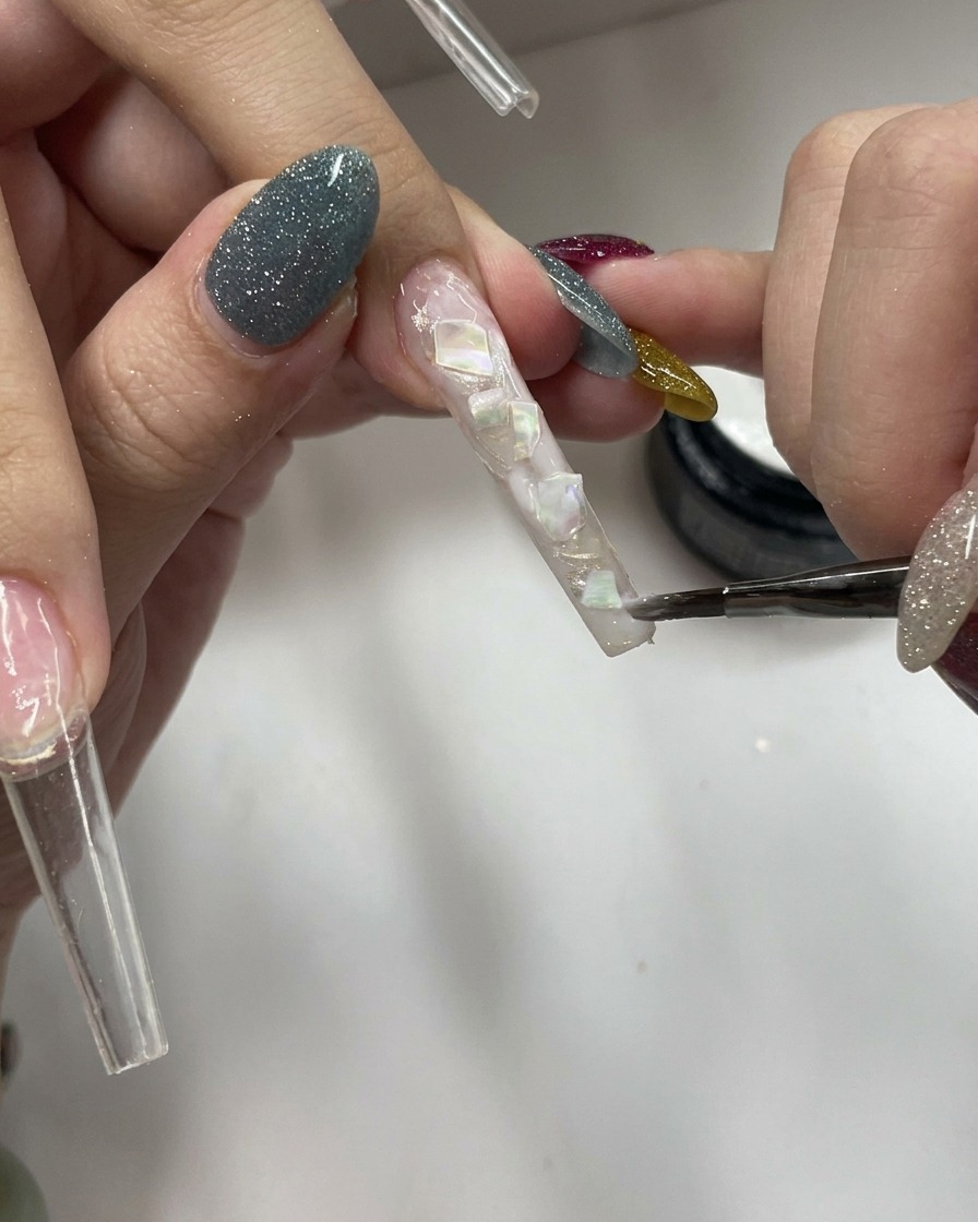 Ein Pinsel trägt eine Schicht transparentes, milchig-weißes Gel auf eine texturierte Nageloberfläche auf.