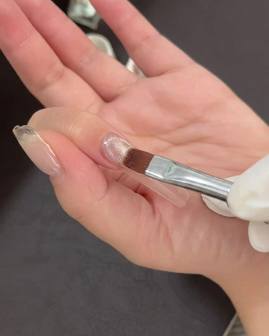 Ein Nailart-Pinsel beim Auftragen einer dünnen Schicht schimmernden Cat-Eye-Gellacks auf einen klaren Nagel-Tip.