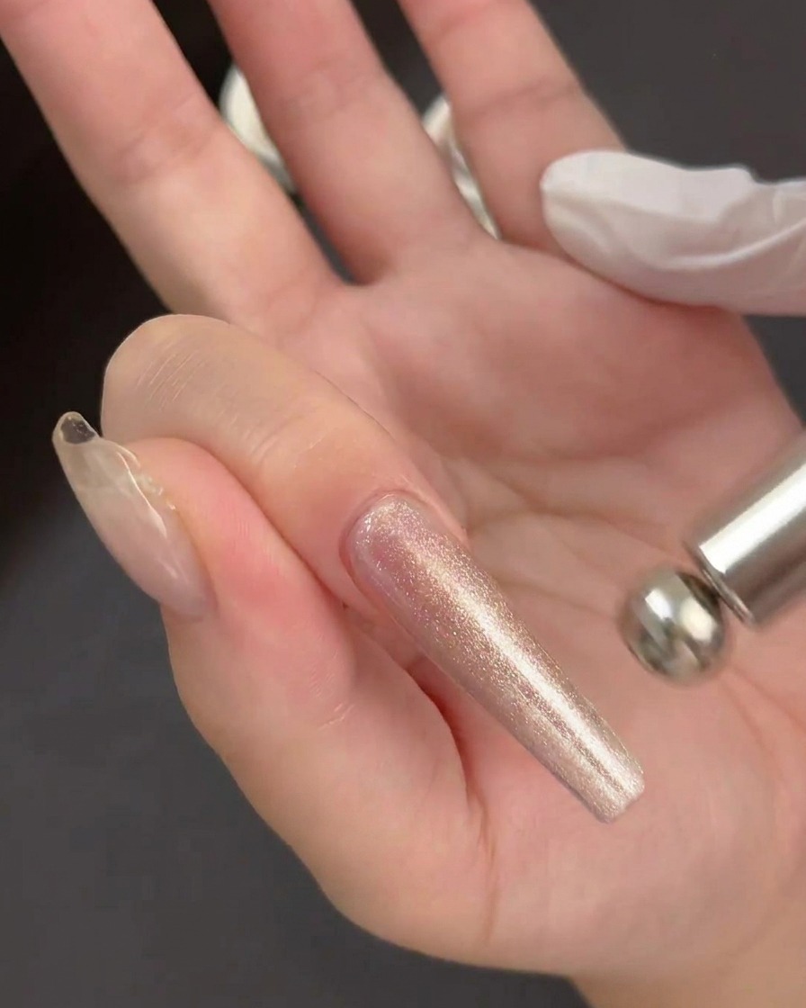 Eine Hand hält ein zylindrisches Magnetwerkzeug direkt über einen nassen, glitzernden Gelauftrag auf einer klaren Nagelverlängerung.