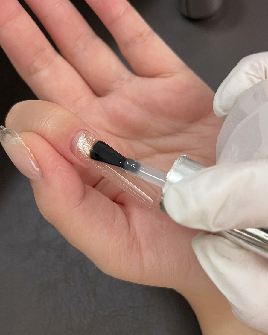 Ein professioneller Nagelpinsel beim Auftragen eines klaren, glänzenden Top Coats über eine schimmernde Cat-Eye-Gelbasis.
