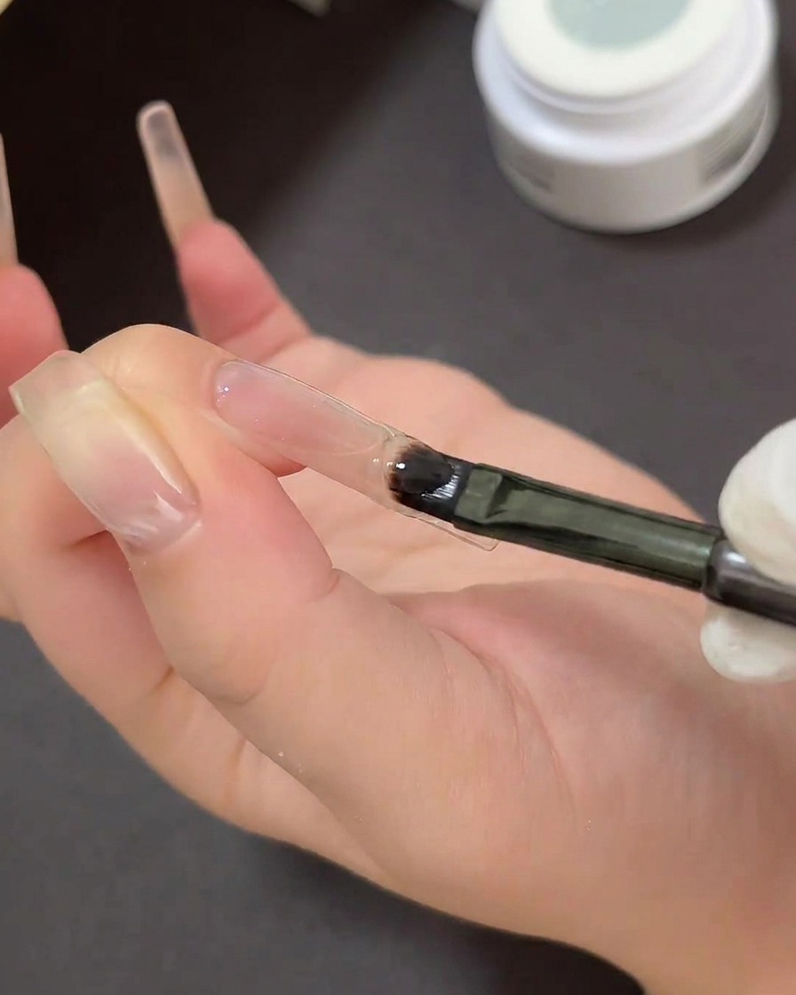 Ein flacher Gelpinsel beim Auftragen von klarem klebrigen Gel auf die Spitze eines Nagels über einer geschwungenen French-Tip-Schablone.