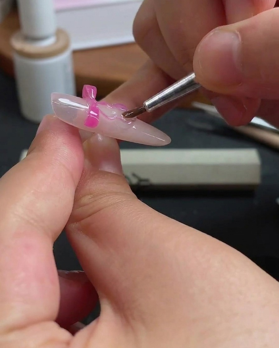 Ein dünner Nagelpinsel trägt vorsichtig klares Gel um die Kanten eines rosa Schmetterlings-Charms auf einem Fingernagel auf.