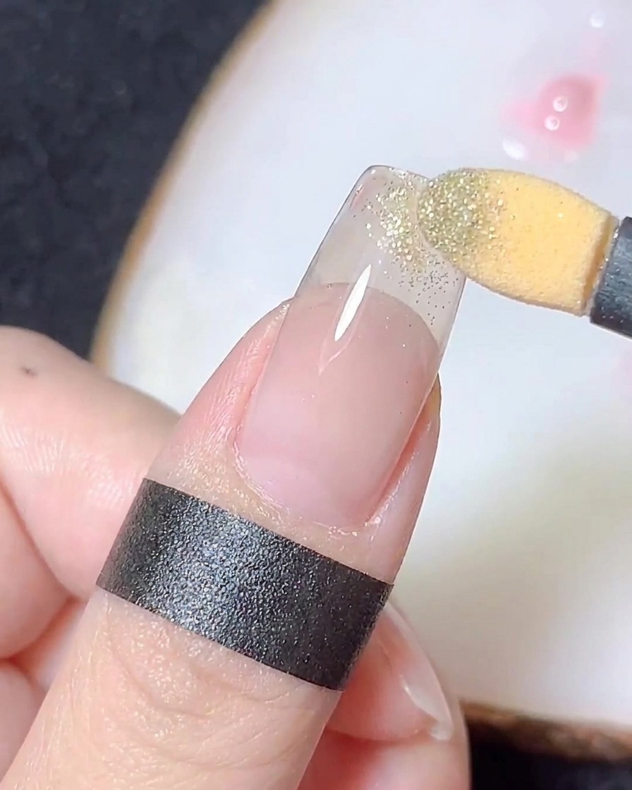 Ein Schwammapplikator reibt goldenes Chrome-Puder auf die Spitze eines klaren Nageltipps, um einen Farbverlauf zu erzeugen.
