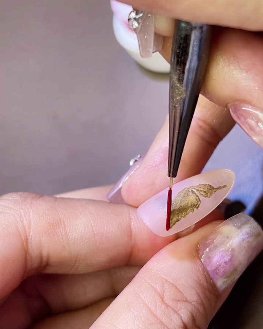 Ein feiner Liner-Pinsel zieht eine dünne rote Gel-Linie um einen goldenen Schmetterlingsflügel auf dem Nagel.