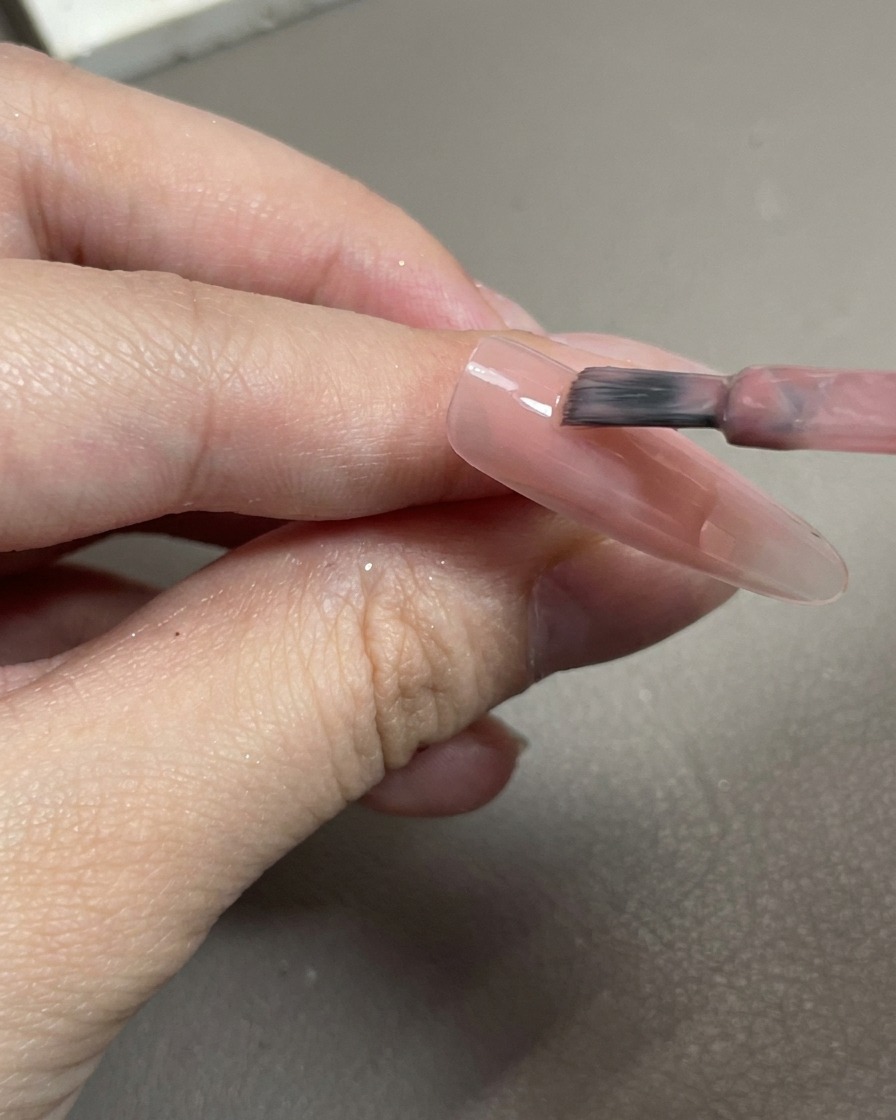 Nahaufnahme eines Pinsels, der einen zartrosa Gellack auf einen durchsichtigen Nageltip aufträgt.