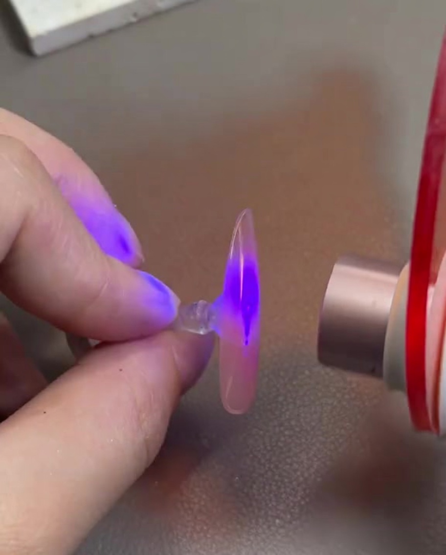 Eine Hand hält einen durchsichtigen Nageltip mit rosafarbenem Gel unter eine kleine UV/LED-Härtungslampe.