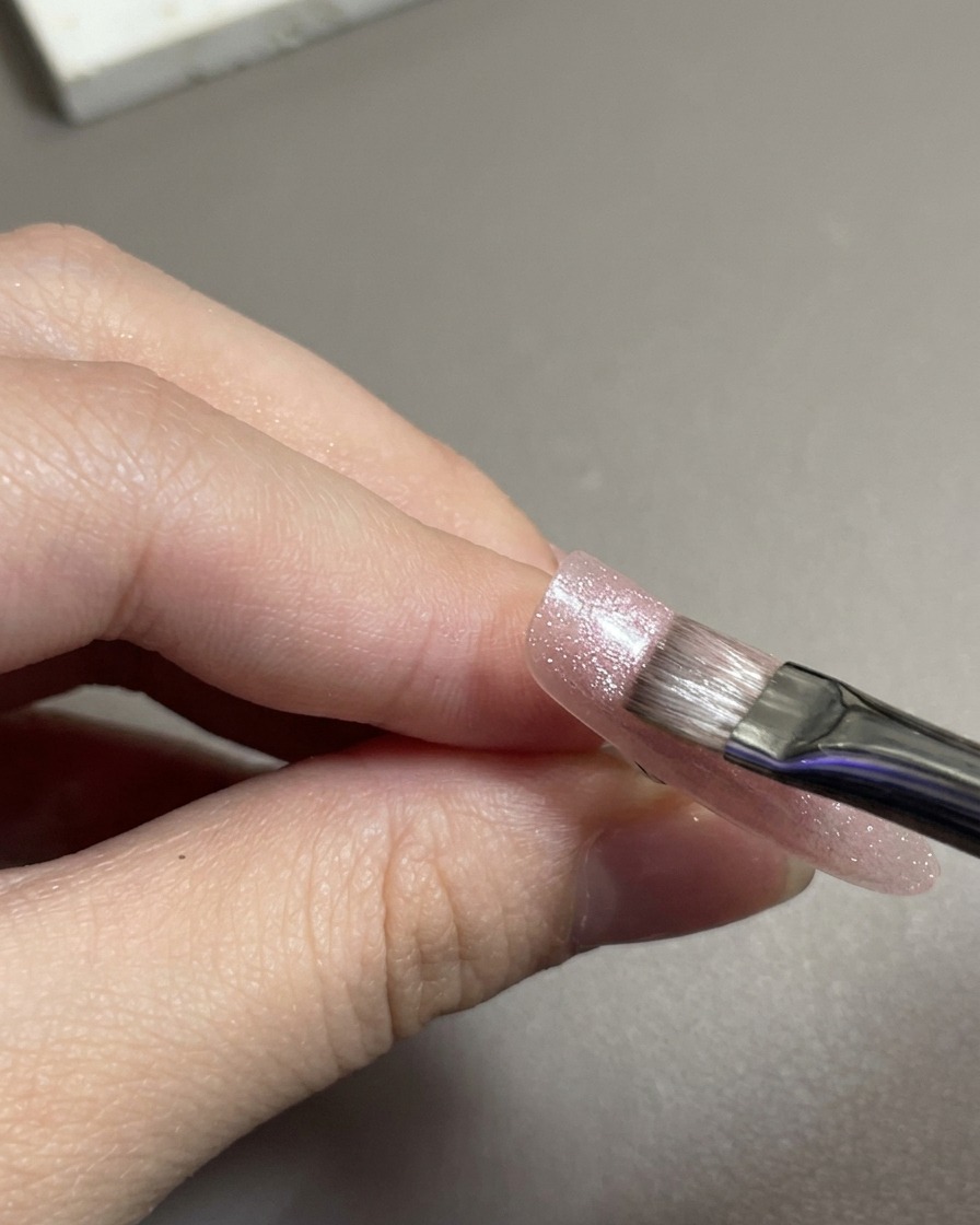 Ein flacher Nagelpinsel verteilt schimmerndes, rosafarbenes Cat-Eye-Magnet-Gel über den Nageltip.