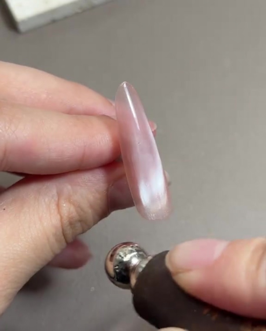 Eine Hand hält ein zylindrisches Magnetwerkzeug nah an einen langen Fingernagel, der mit zartrosa feuchtem Cat-Eye-Gel lackiert ist, und verschiebt die Magnetpartikel.