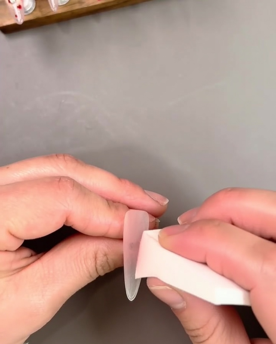 Hand hält eine Nagelmodellage, während ein transparenter rosa Gel-Lack mit einem Pinsel aufgetragen wird.