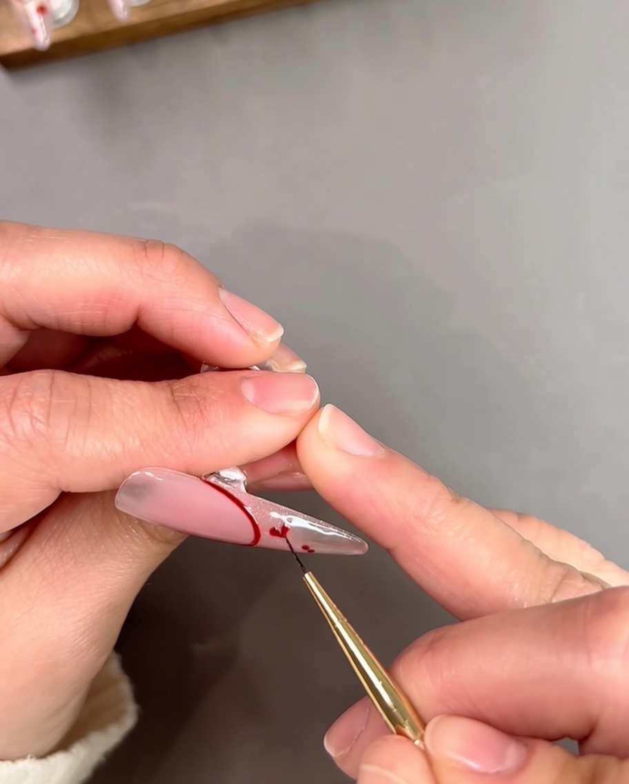 Nahaufnahme einer Nageldesignerin, die mit einem dünnen Pinsel zwei kleine rote Punkte auf einem klaren Nagel zu einem Herz verbindet.
