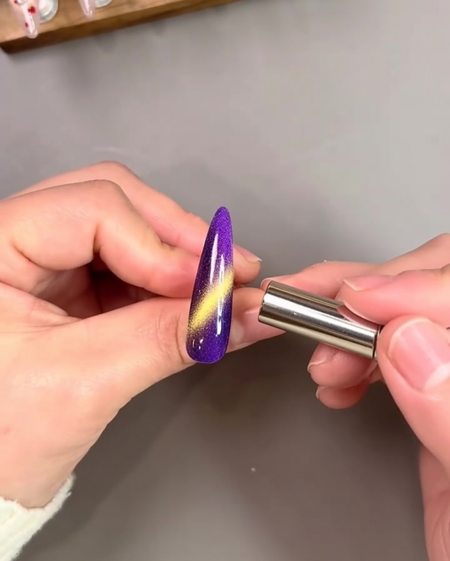 Eine Hand hält einen zylindrischen Magneten nahe an einen leuchtend violett lackierten Nagel, wodurch ein heller goldener Cat-Eye-Streifen aktiviert wird.