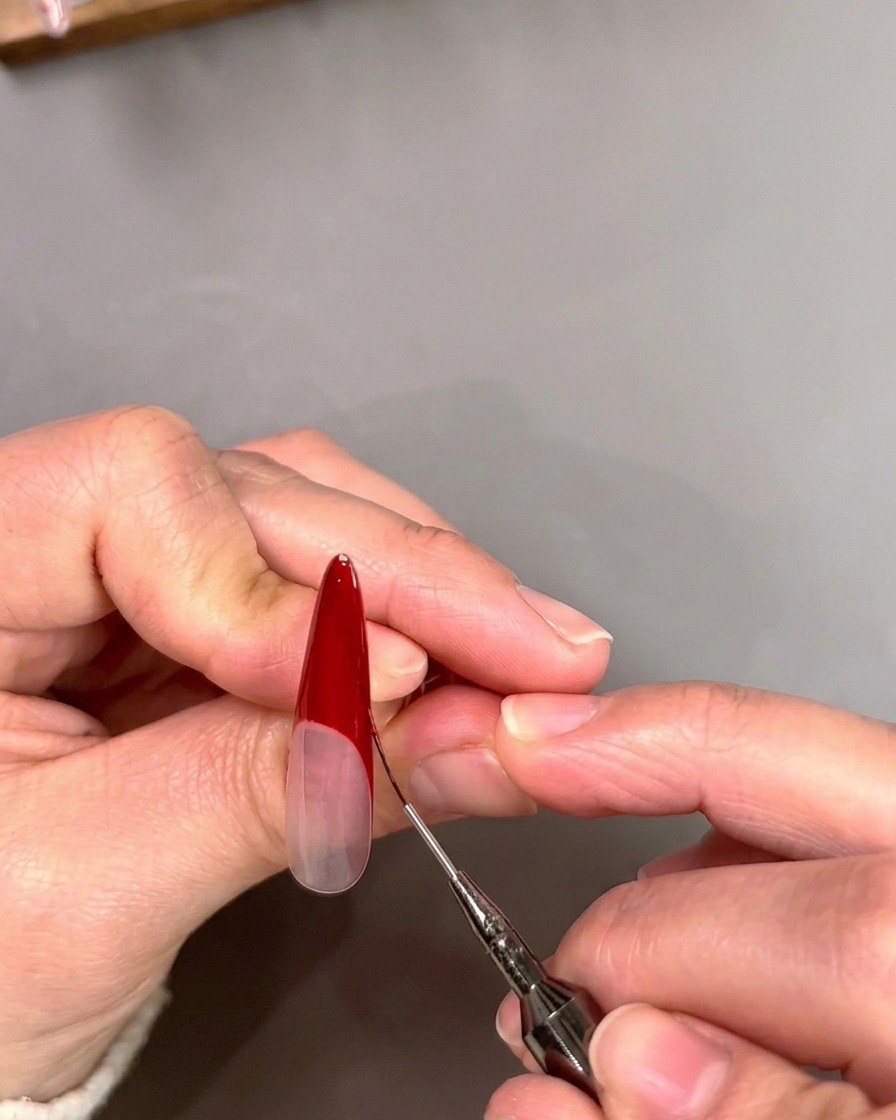 Ein dünner Liner-Pinsel schärft die innere Kante einer tiefroten, V-förmigen French-Spitze auf einem Nagel.