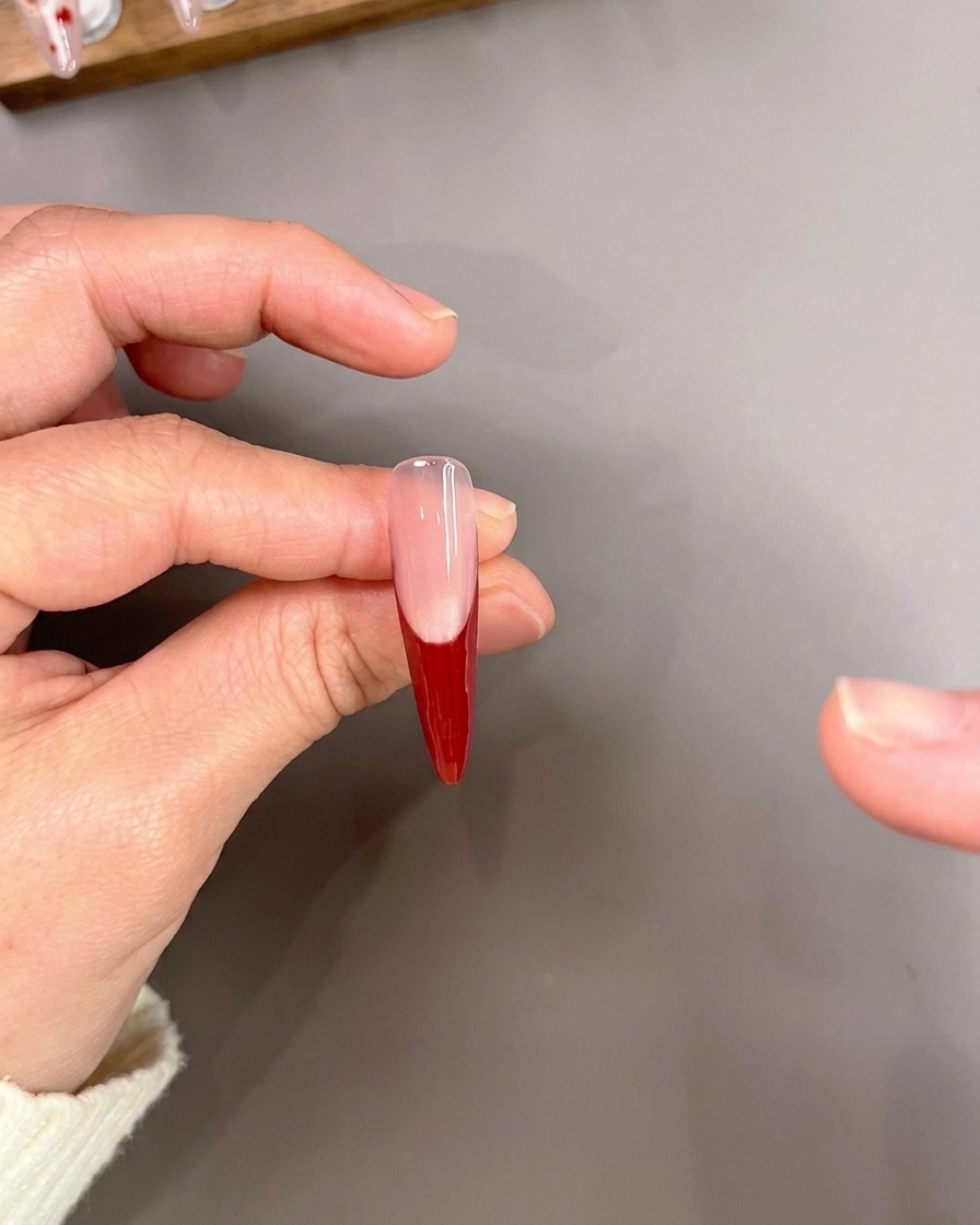 Ein Liner-Pinsel zeichnet vorsichtig dünne rote Linien auf eine Nagelspitze mit einer roten French-Maniküre-Basis.