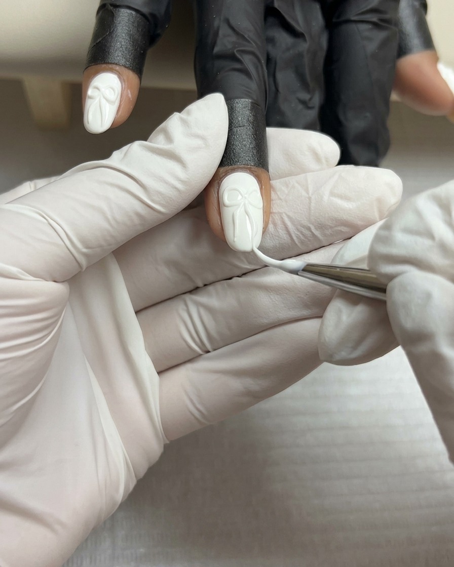 Eine Nahaufnahme eines feinen Liner-Pinsels, der dünne weiße Linien auf die Oberfläche einer 3D-Gelschleife auf einem weißen Nagel zeichnet.