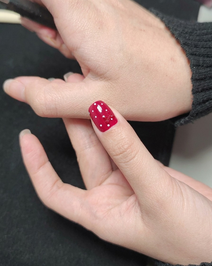 Ein Nagellackpinsel trägt einen glänzenden klaren Top Coat über einen roten Nagel mit weißen Punkten und einem roten Schleifen-Charm auf.