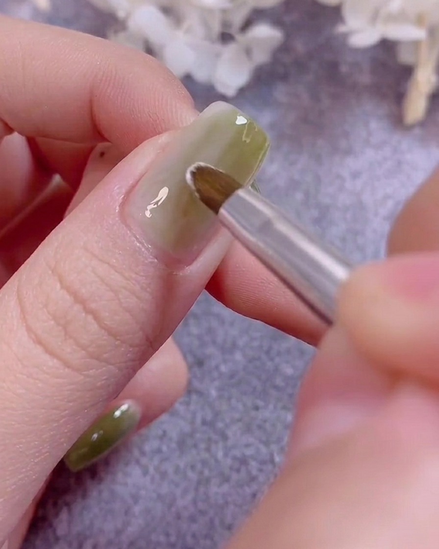 Ein feiner Nailart-Pinsel malt zarte weiße Blütenblätter auf einen grün lackierten Nagel.
