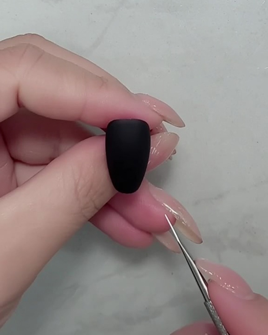 Nahaufnahme eines Nagelpinsels beim Malen dünner weißer Stiele auf einem mattschwarzen Nagel.