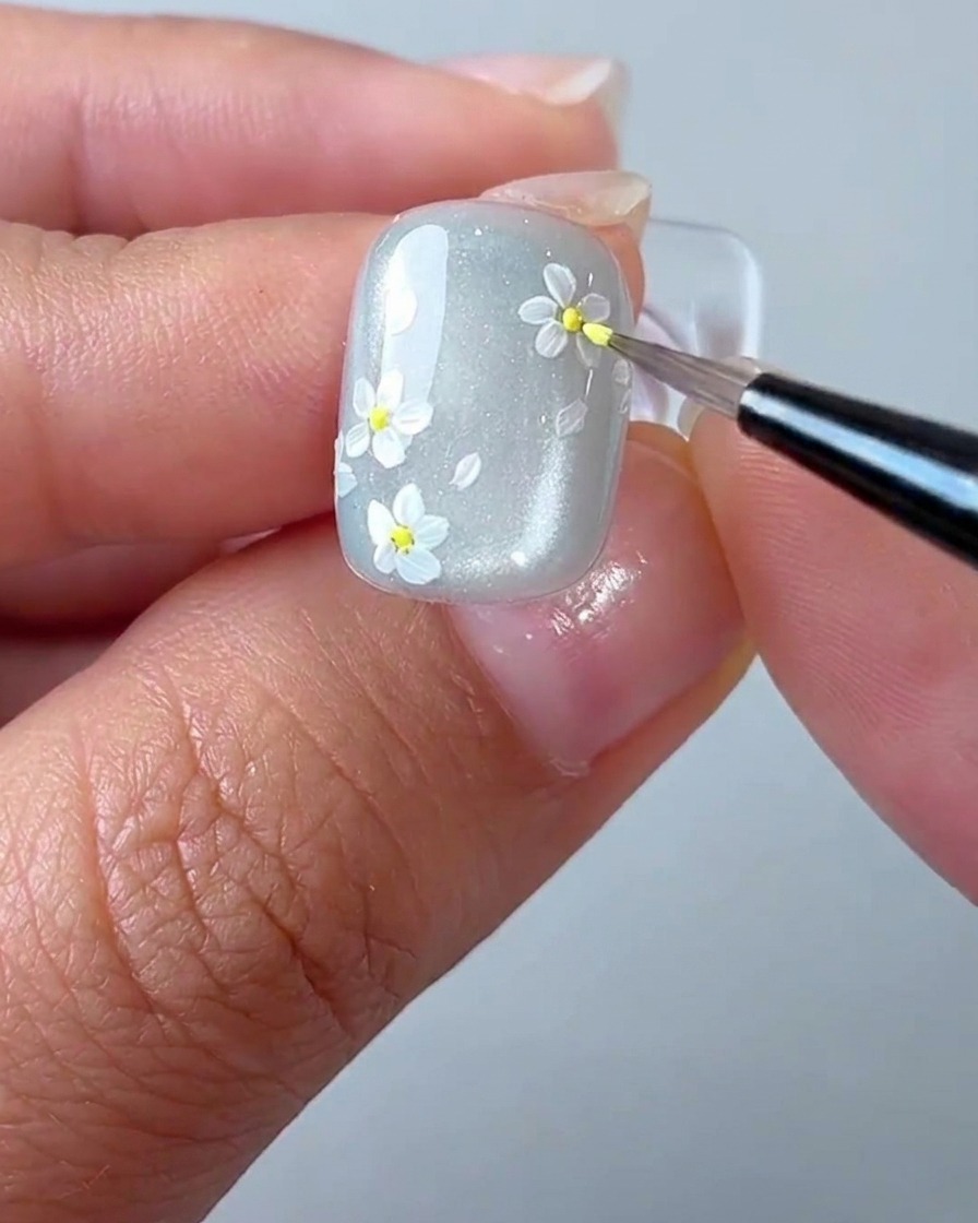 Ein Detail-Pinsel setzt einen winzigen gelben Punkt in die Mitte eines weißen Gänseblümchens auf dem Nagel.